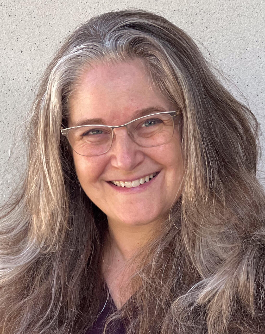 White middle-aged woman wearing yellow metal half-spectacles with brown flowing hair to below her shoulders smiles, eye crinkling. She is wearing a merlot coloured tunic.